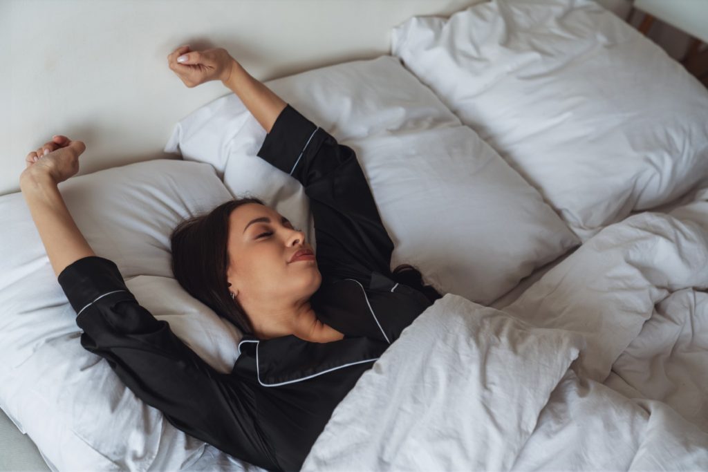 A young beautiful woman has just woken up after sleeping in bed and stretches her arms up in her pajamas under a blanket in her bedroom.